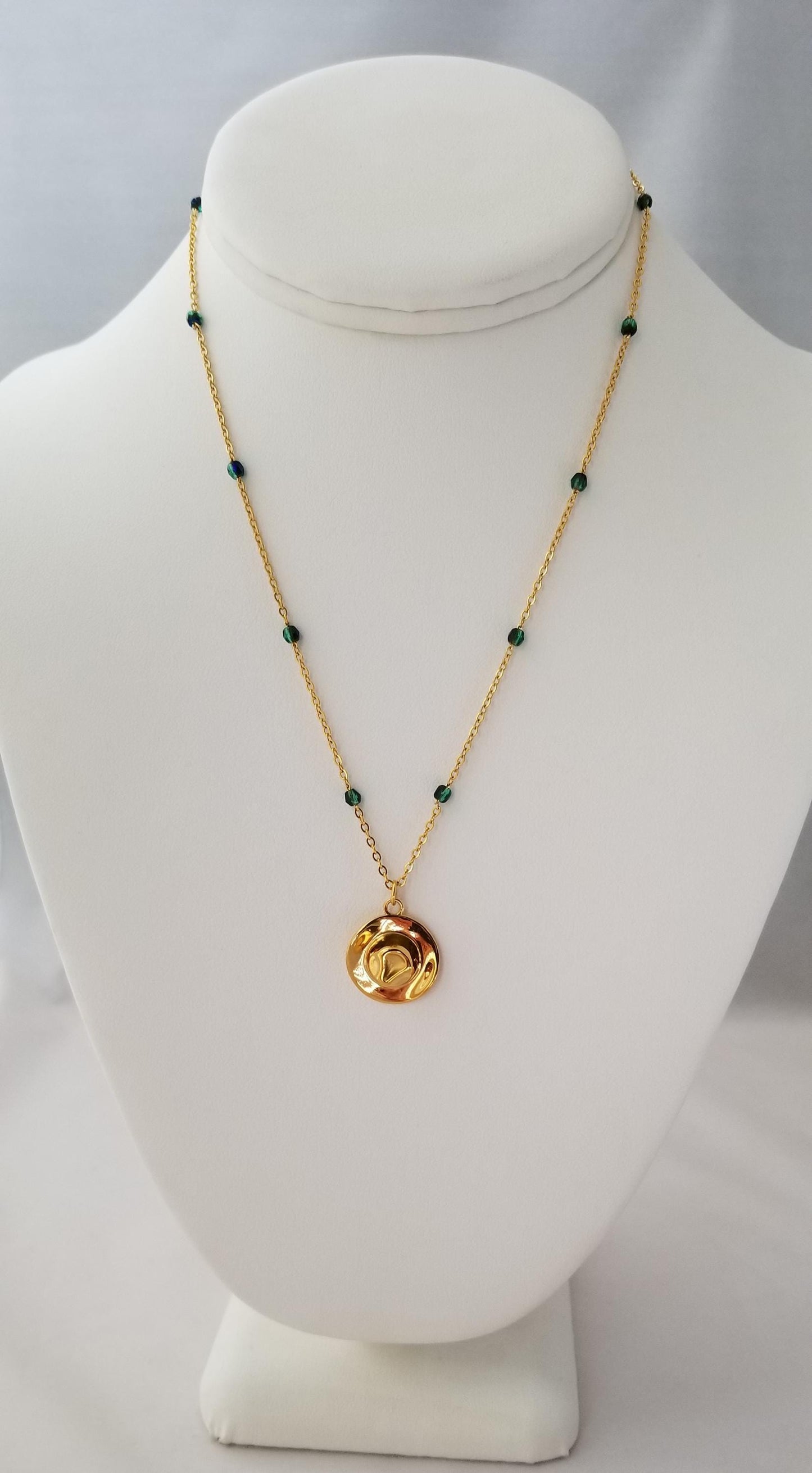 A gold necklace with green beads and a round pendant is displayed on a white bust form.