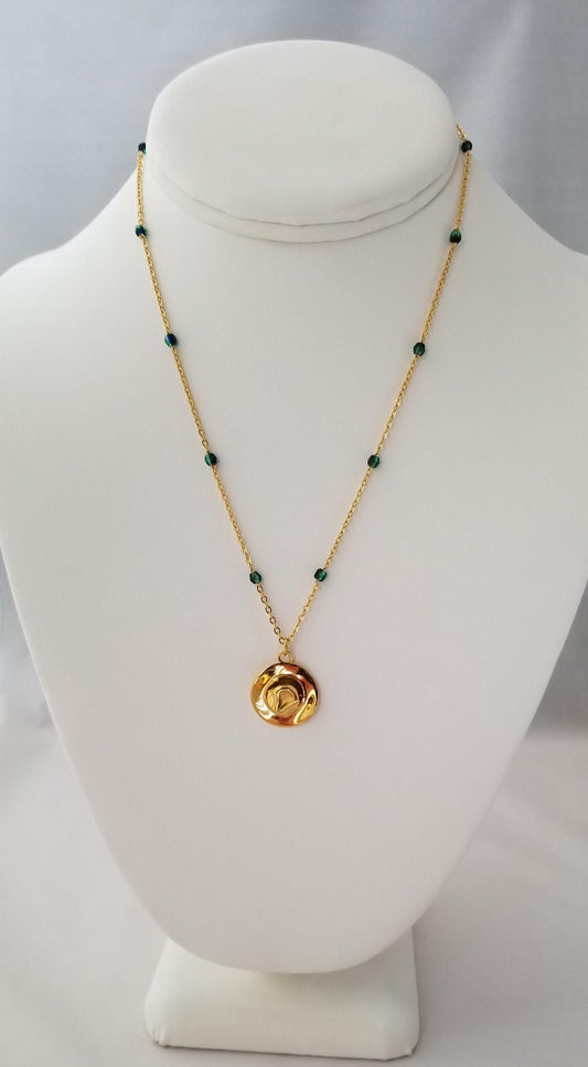 A gold necklace with green beads and a round pendant is displayed on a white bust form.